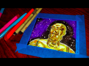 C3PO the robot from Star Wars is looking into a starry night in a small drawing framed by blue tape. Pens used to draw it are next to the drawing.
