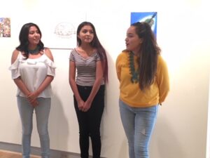 Three teenaged girls dressed casually standing in front of a white wall with a blue painting hanging behind them