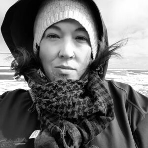 A woman with dark eyes and a tattooed chin stands in an icy landscape wearing a coat, scarf, and knitted cap